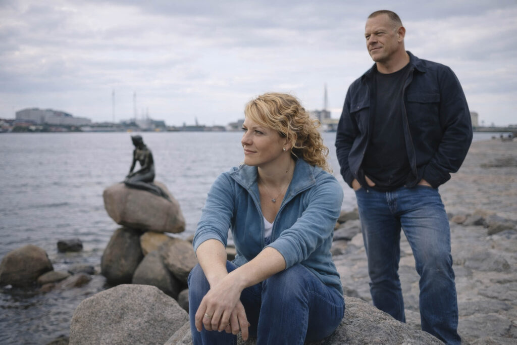 June Haunsby seated by the water with Dane Haunsby standing behind her near the shoreline Little Mermaid