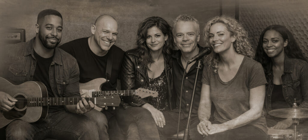 Black-and-white group portrait of Luca, Dane, Nikki, Mekel, June, and Zaya Haunsby in a relaxed rehearsal studio with guitars, microphone, and drums.